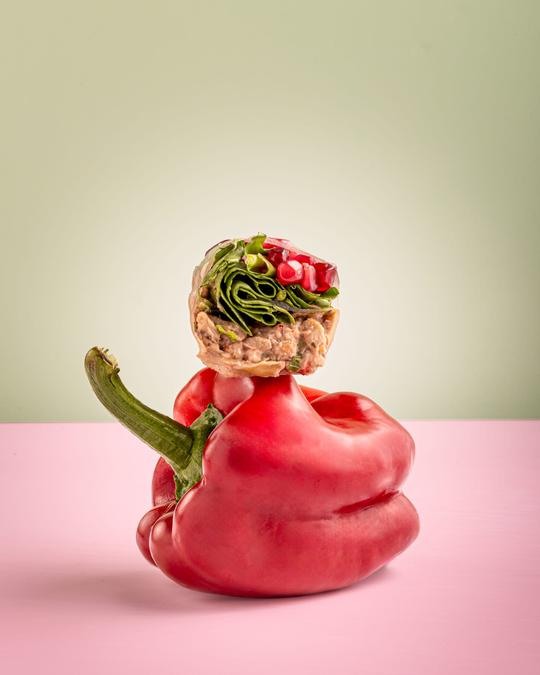 A vine leaf and rice roll balanced upright on a red pepper against a pink surface and pale sage ground