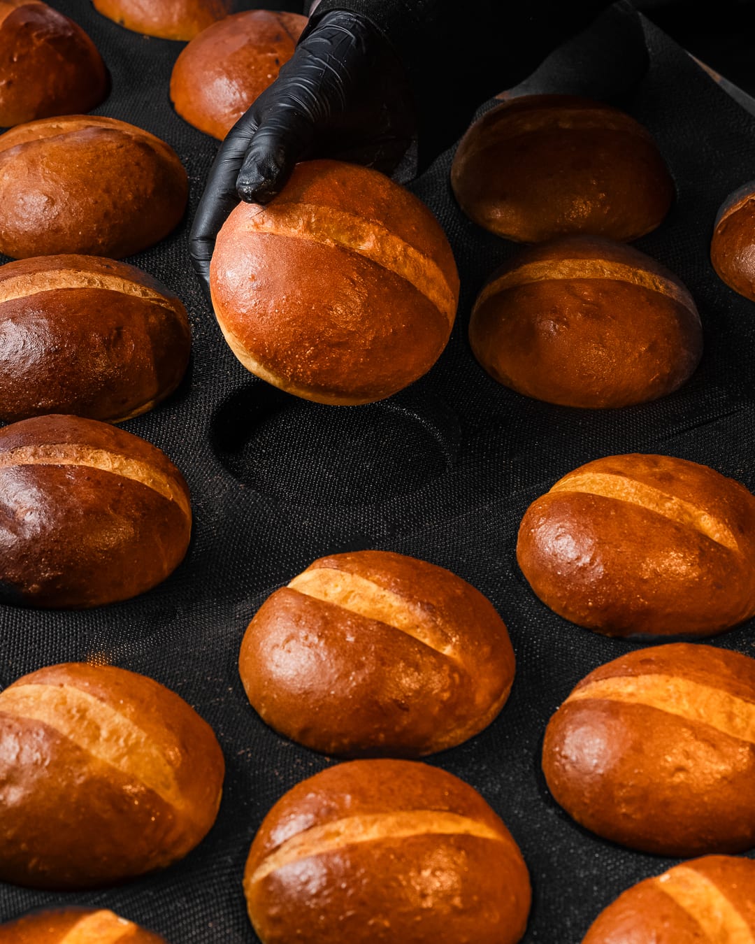 A gloved hand setting a glossy brioche bun among rows of buns on a dark tray