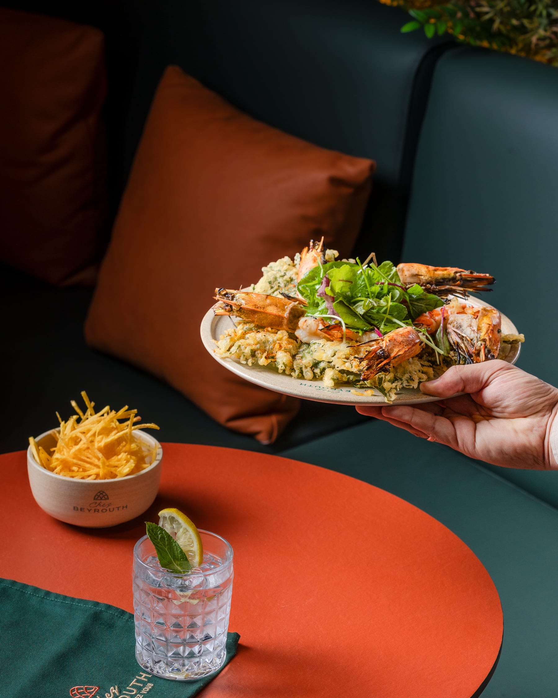 A hand holding a plate of prawns and tempura above an orange table set with a cocktail and a bowl of fries