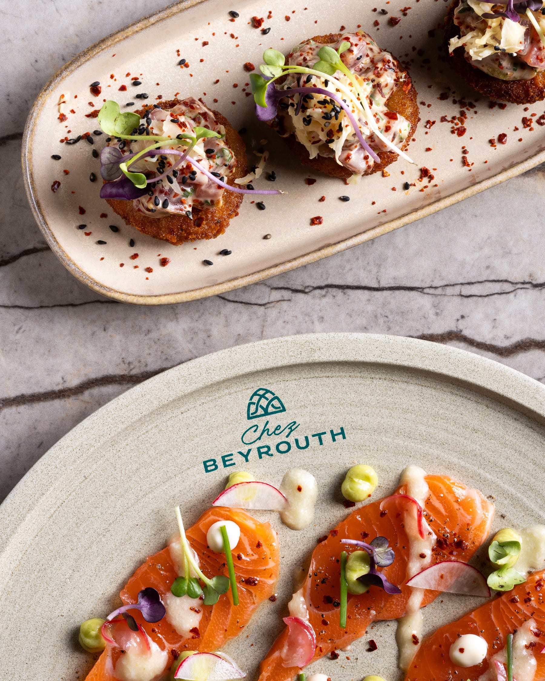 An overhead of canapés on an oval plate beside salmon crudo on a Chez Beyrouth plate, on marble