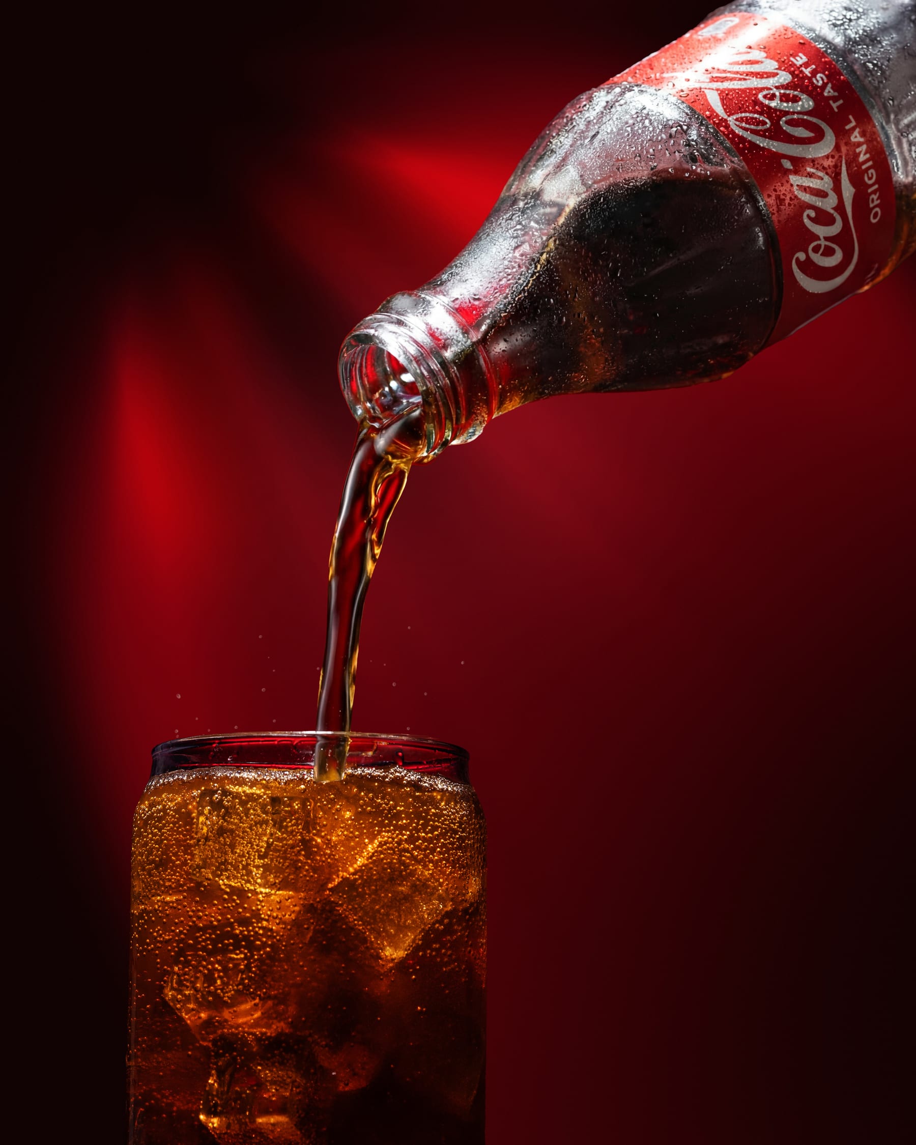 Coca-Cola poured from a glass bottle into a tumbler of ice against a deep red ground