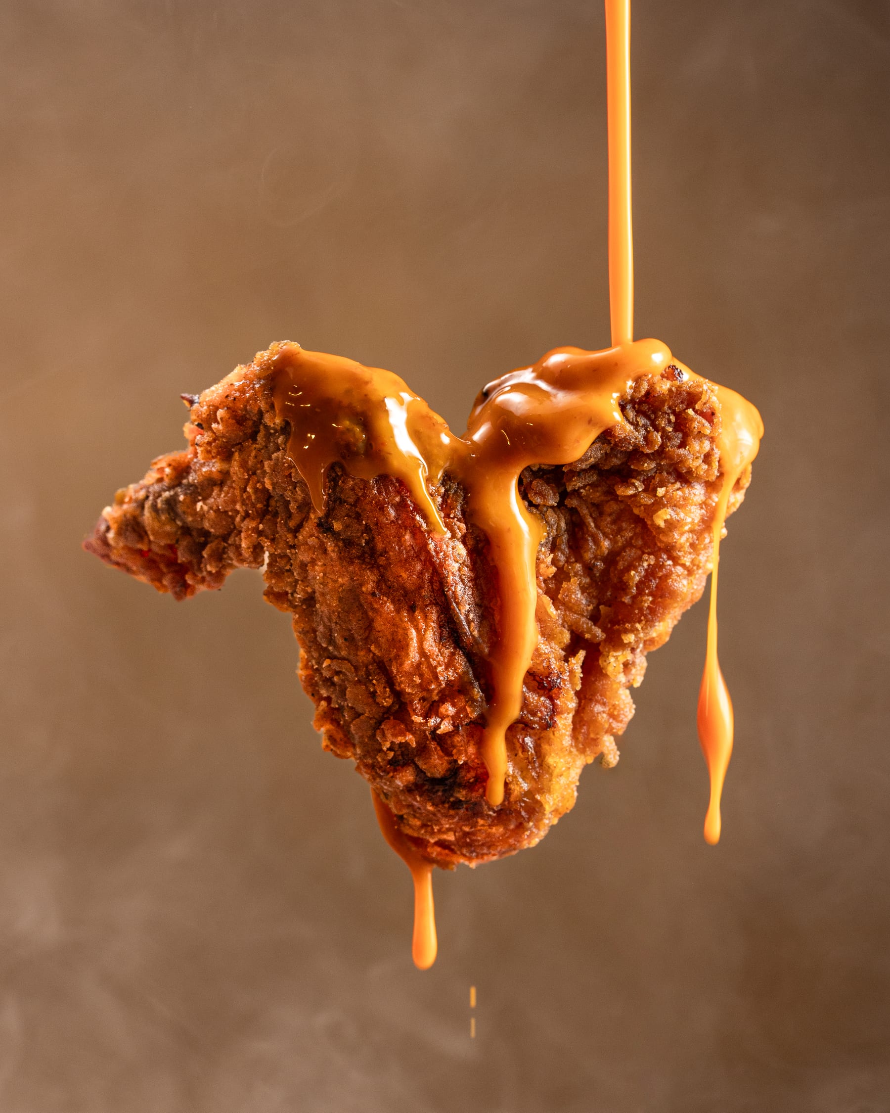 A craggy piece of fried chicken held in mid-air with sauce running off it against a warm brown ground