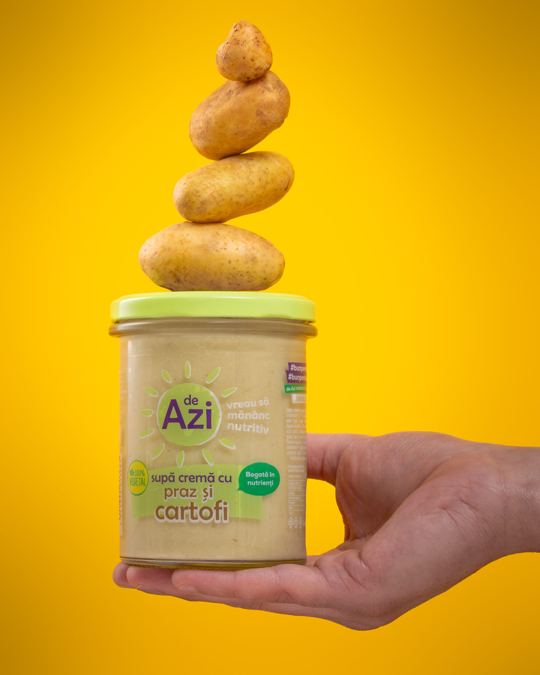 A hand holding a de Azi potato and leek soup jar with four potatoes balanced on its lid against yellow