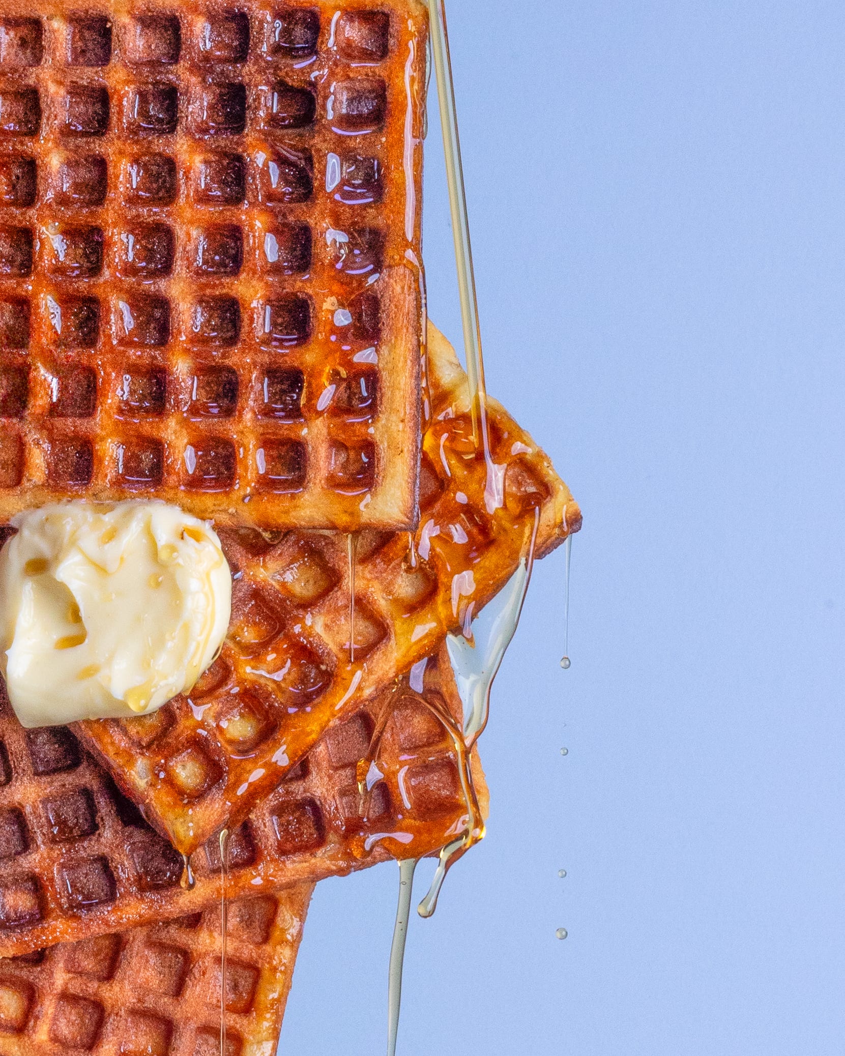 A stack of waffles with a melting knob of butter and honey pouring over the edge against pale blue