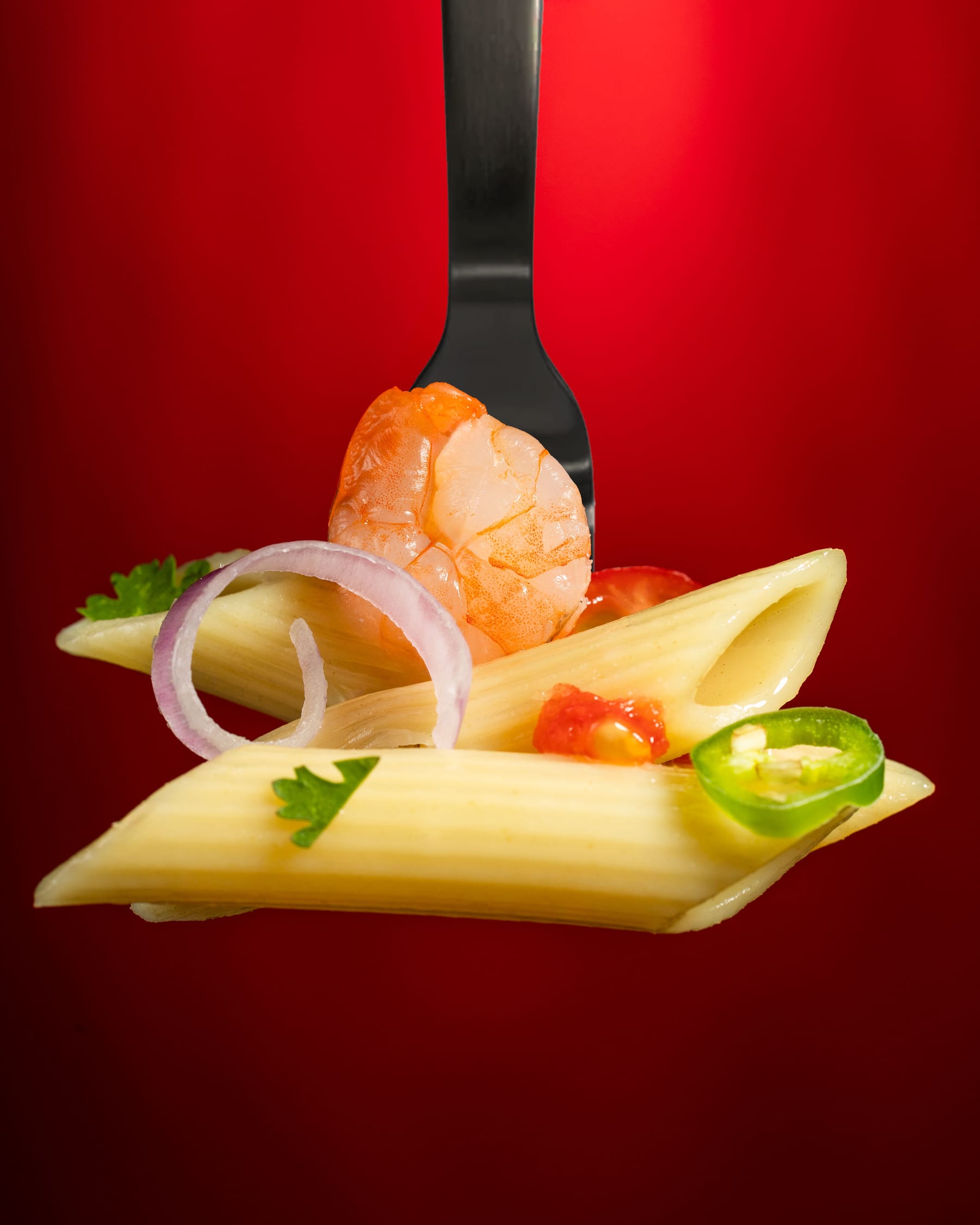 Penne, a prawn, red onion, and a slice of jalapeño balanced on a fork against a red ground