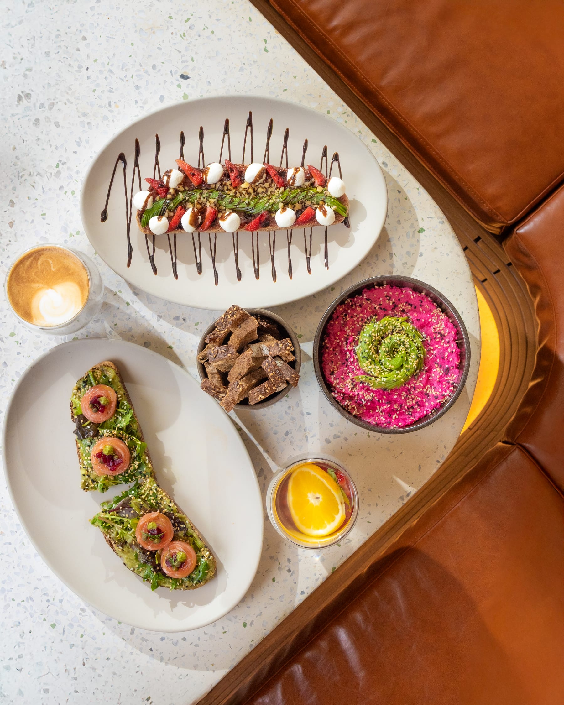 An overhead brunch spread of avocado toast, berry toast, and coffee on a terrazzo table beside a tan leather banquette
