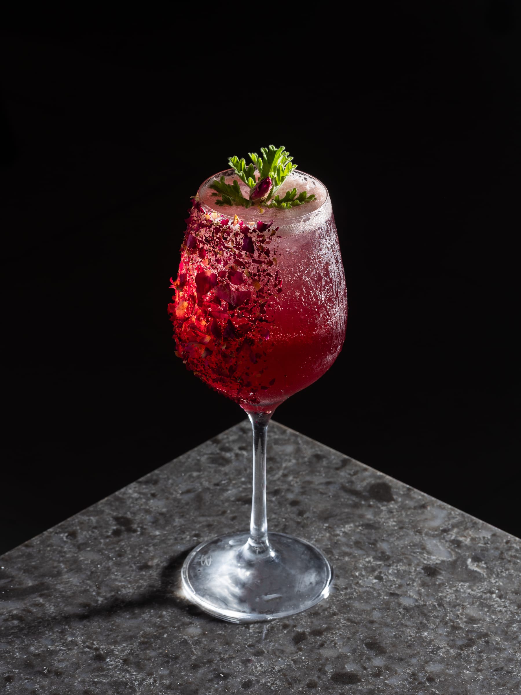 A tall stemmed glass of a red berry cocktail on a granite plinth against black