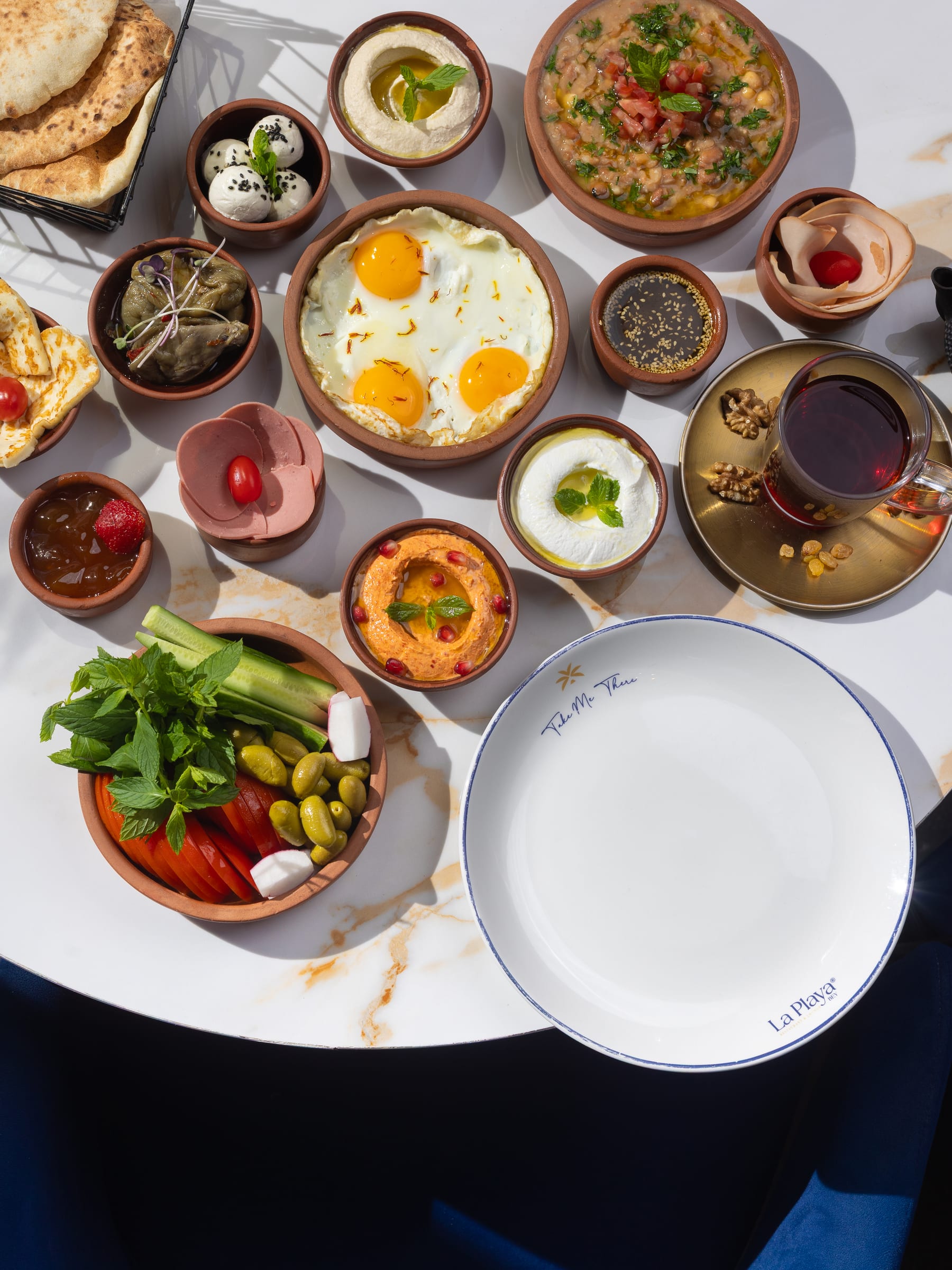 An overhead Lebanese breakfast spread of fried eggs, mezze bowls, tea, and vegetables around a La Playa plate