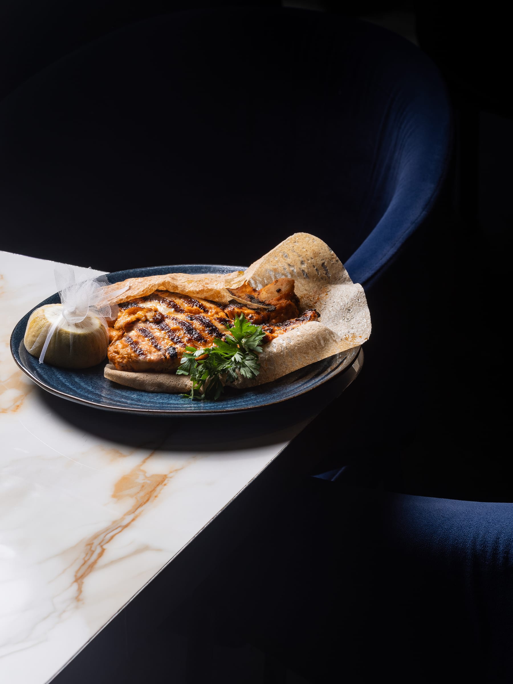 A grilled fish fillet with flatbread on a blue plate at the edge of a marble counter in low light
