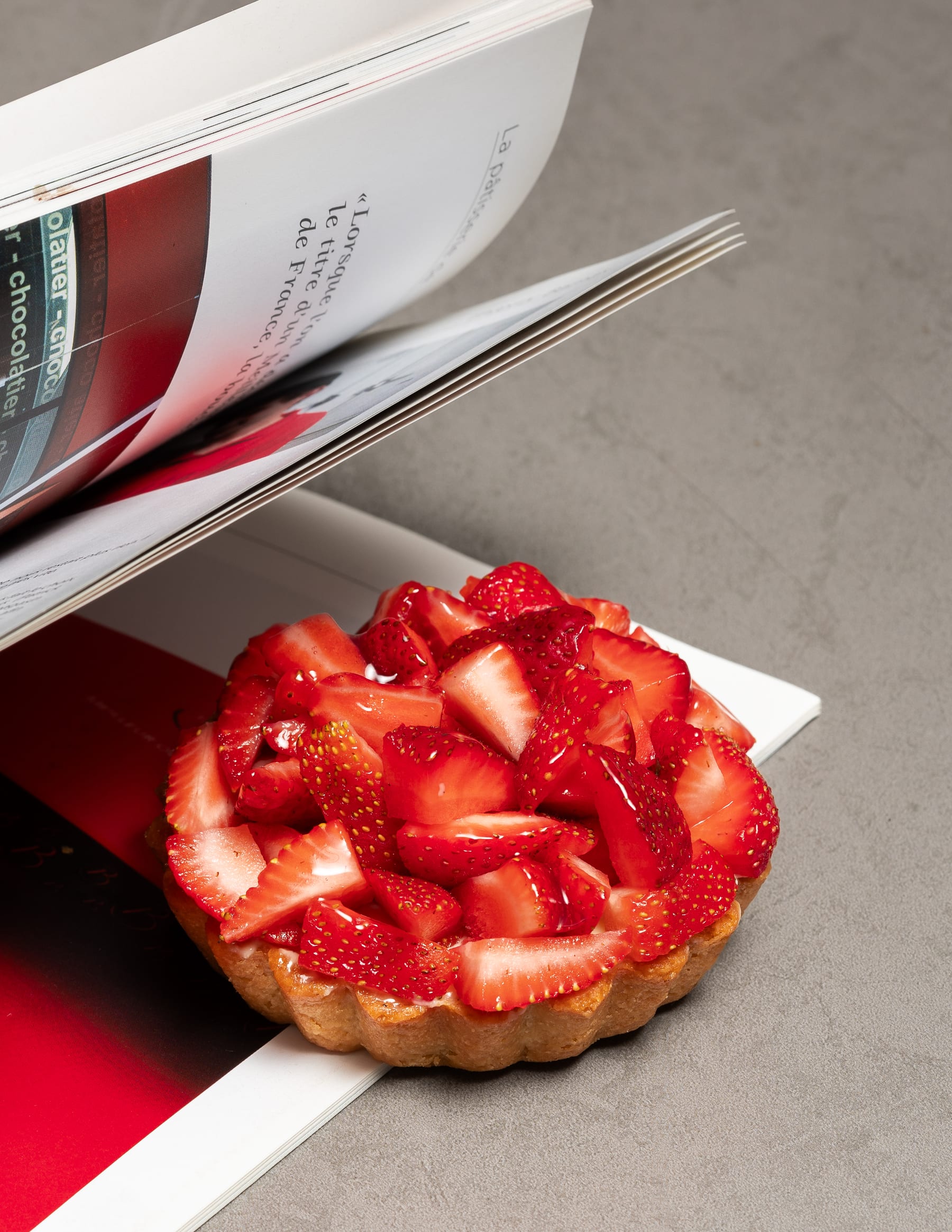 A strawberry tart on a white board with an open chocolate reference book behind it