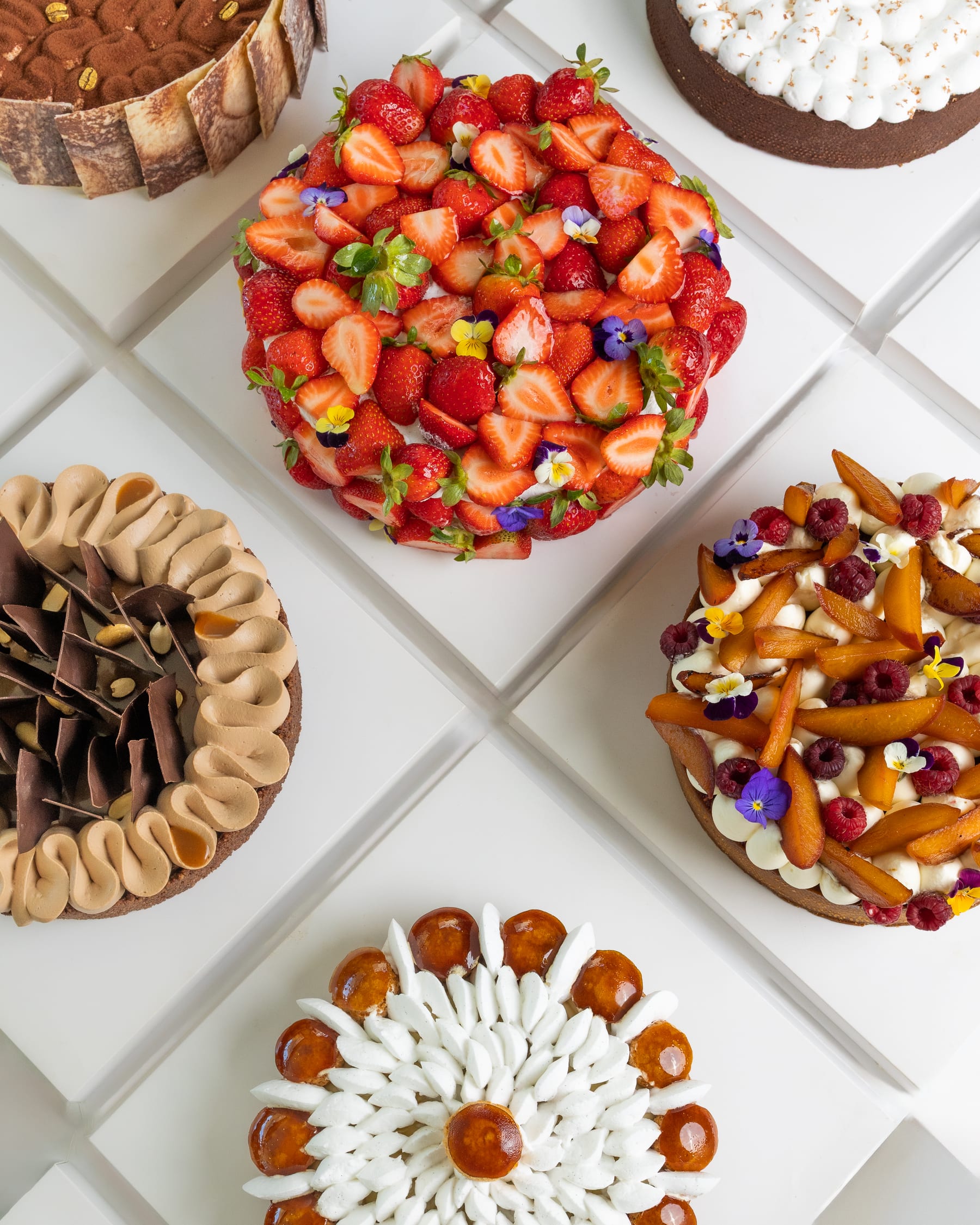 An overhead arrangement of patisserie tarts on white tile — strawberry, chocolate, raspberry, and lemon meringue