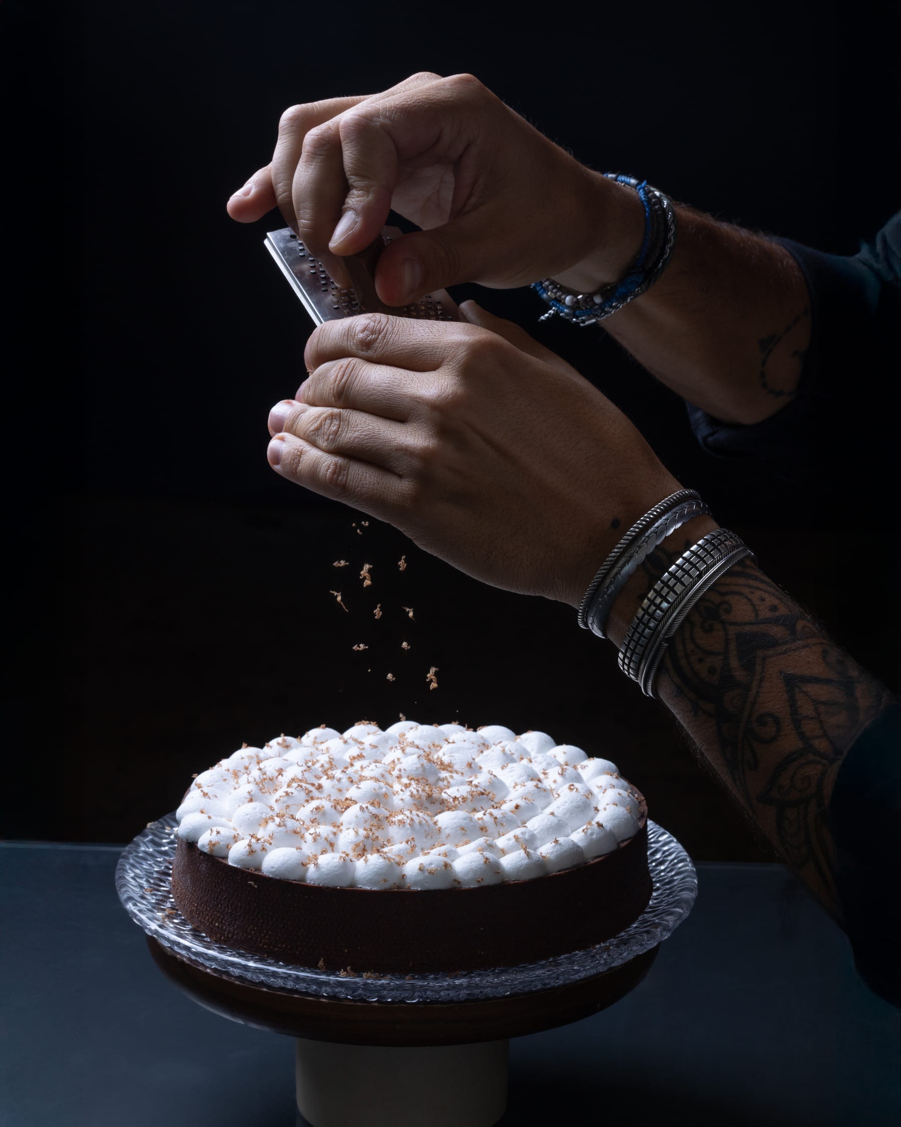 Two hands grating chocolate over a white piped cake on a stand in low light