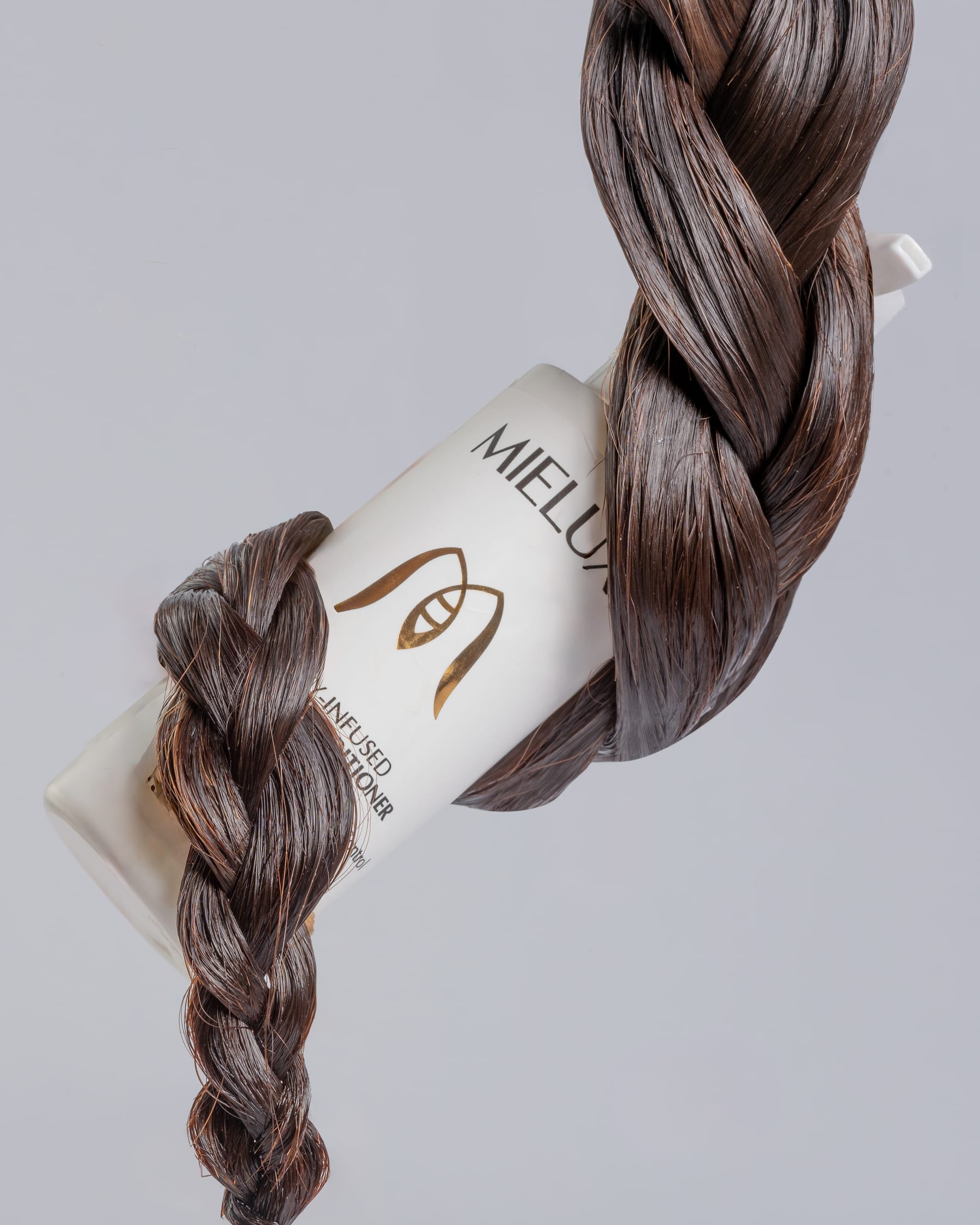 A braid of dark hair wound around a Mielux conditioner bottle against a pale grey ground