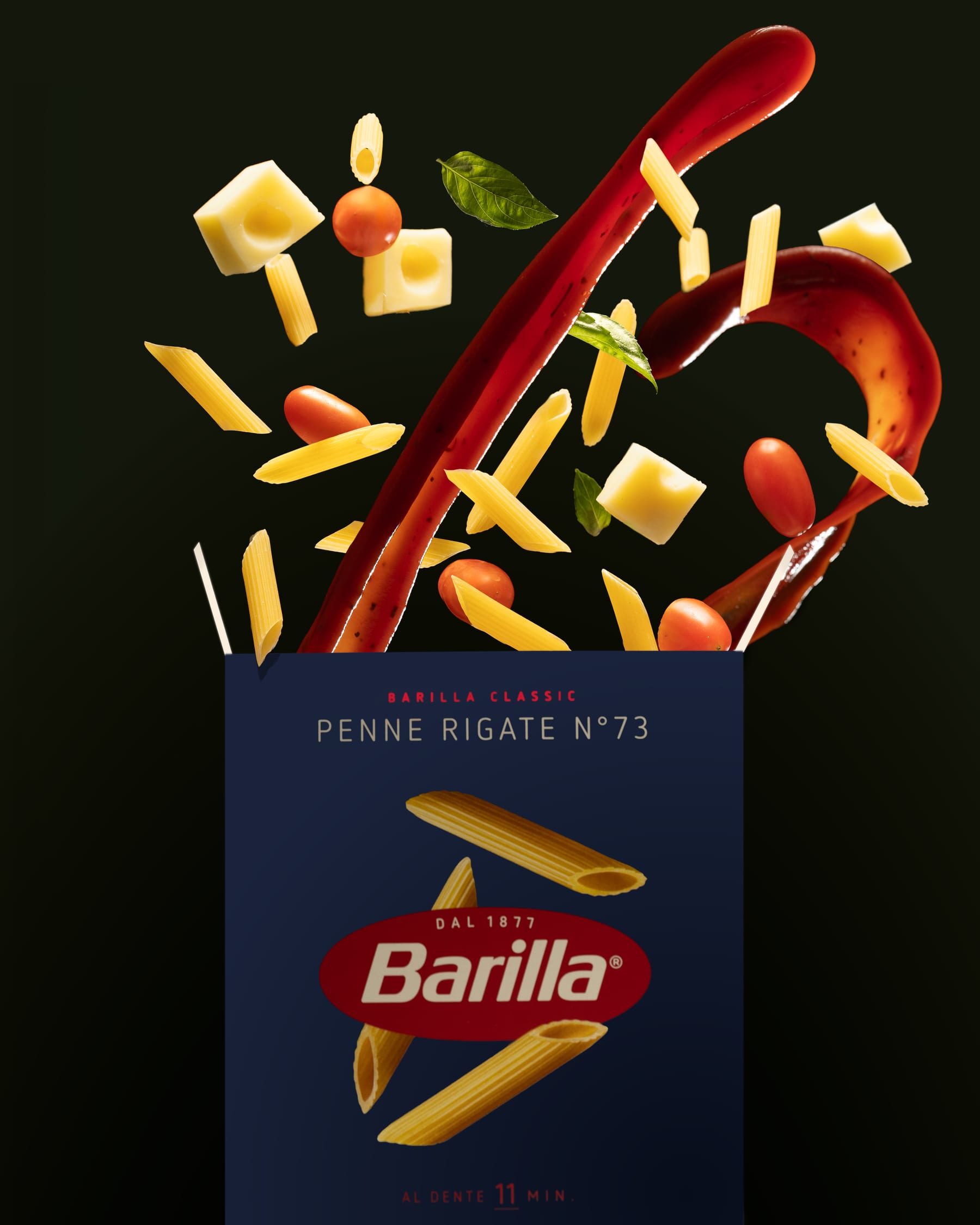 Penne, cheese cubes, tomatoes, and a ribbon of red sauce suspended above a Barilla Penne Rigate box on dark green
