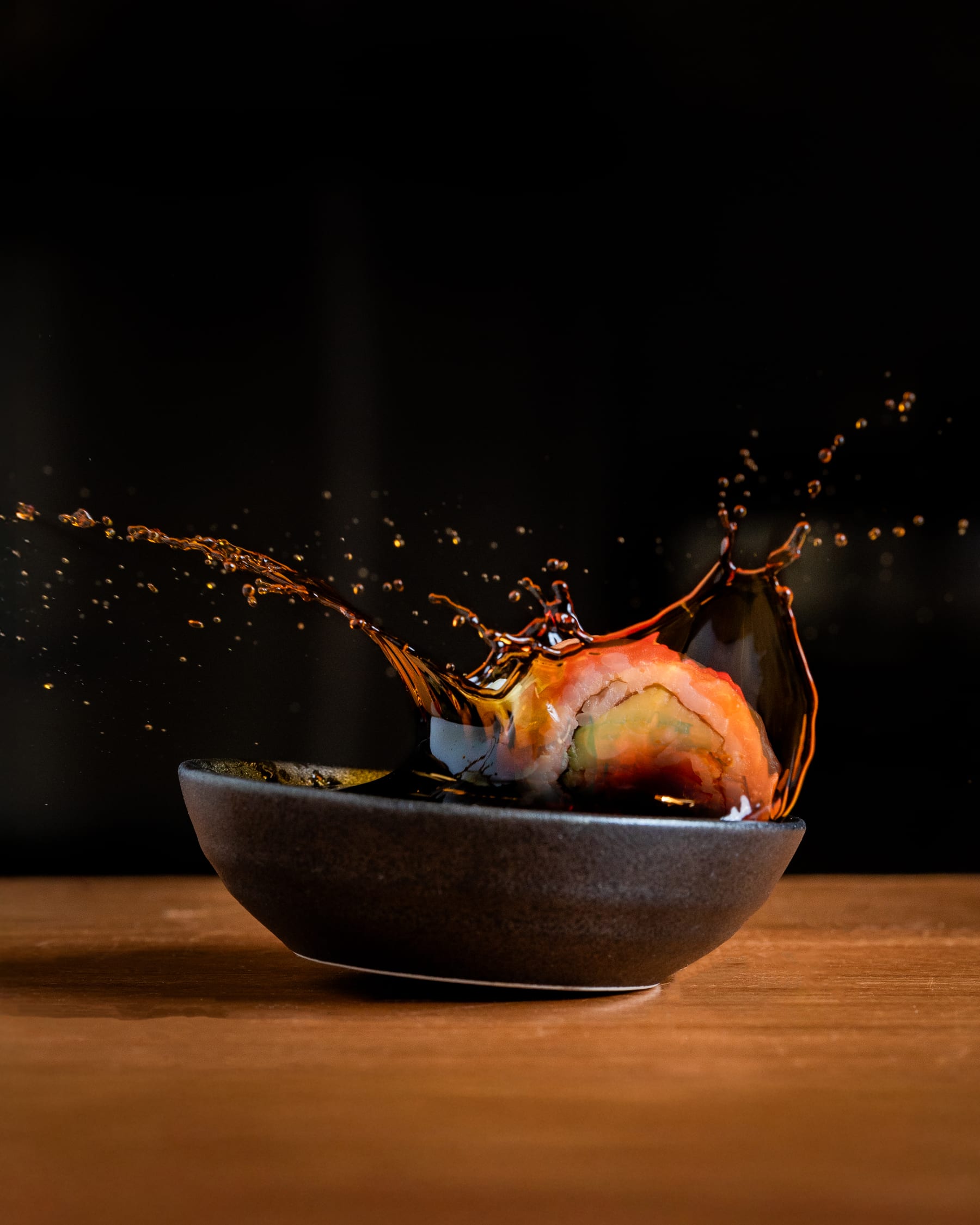 A sushi roll dropping into a dark bowl with sauce splashing up around it