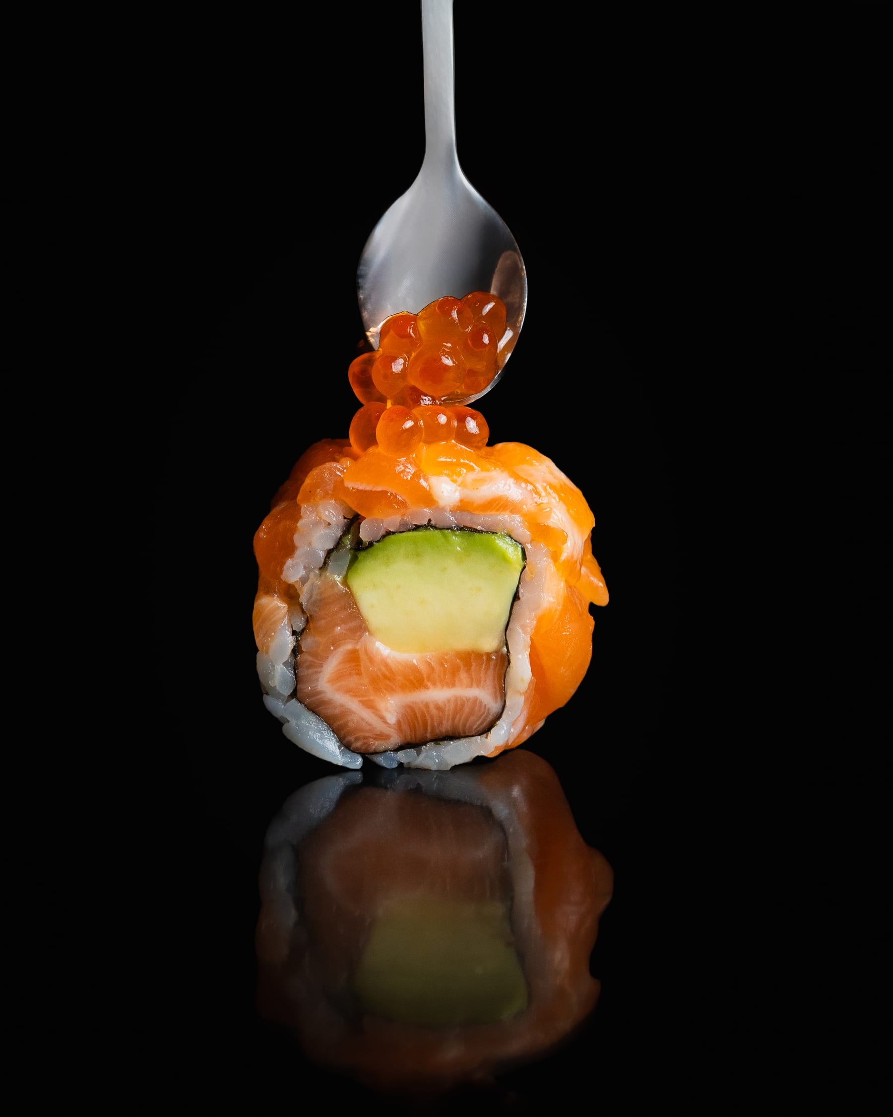 A salmon-wrapped sushi roll topped with roe, a spoon poised above it, on a black reflective surface
