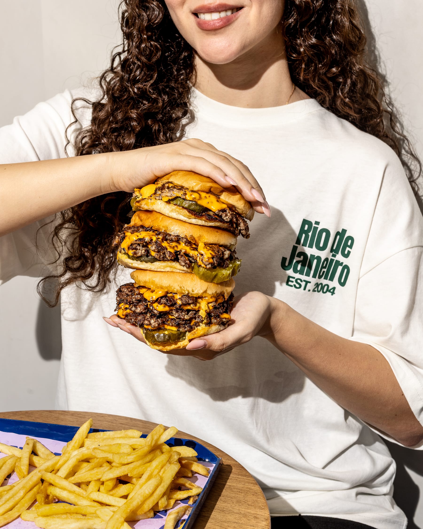 A woman in a white sweatshirt holding a tall stack of smash burgers above a tray of fries