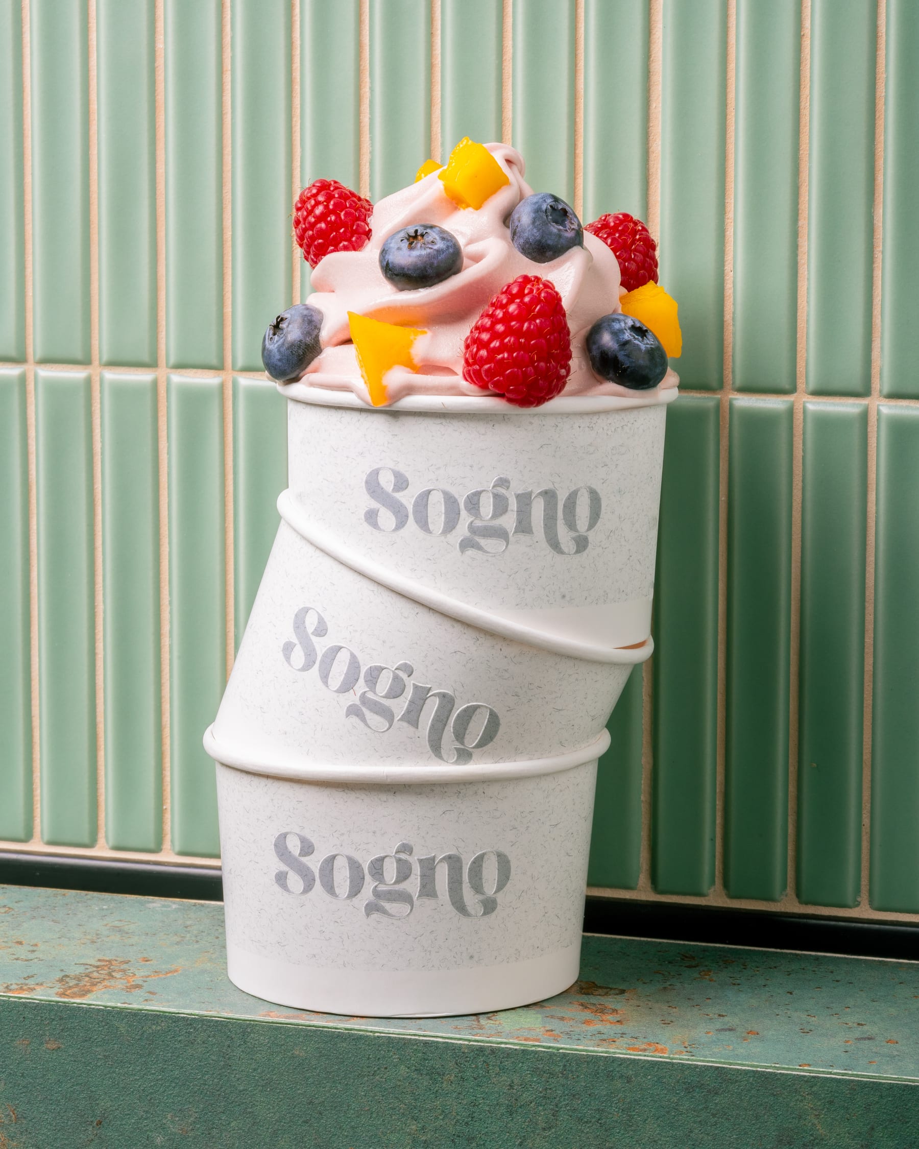 Three stacked Sogno frozen yogurt cups topped with raspberries, blueberries, and mango against green tile