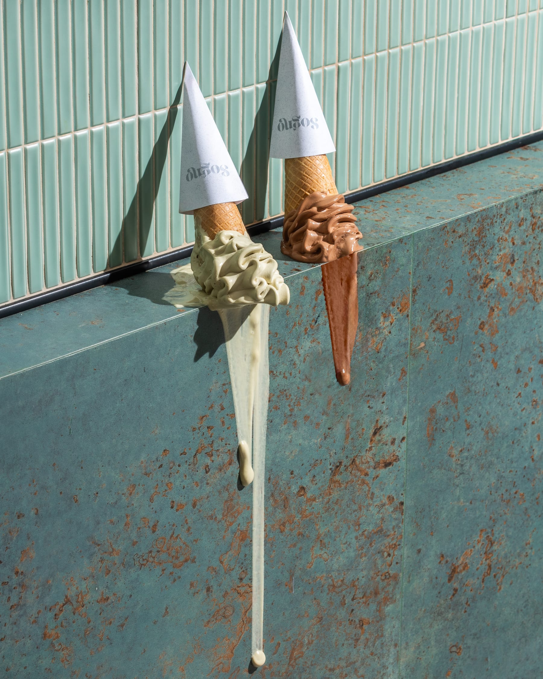 Two melting gelato cones propped against a weathered teal wall with drips running down it