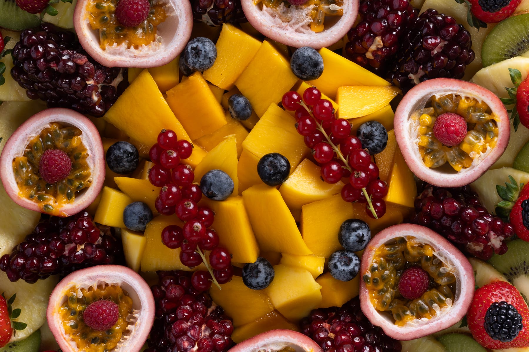 An overhead mosaic of mango cubes, halved passionfruit, pomegranate, blueberries, and redcurrants