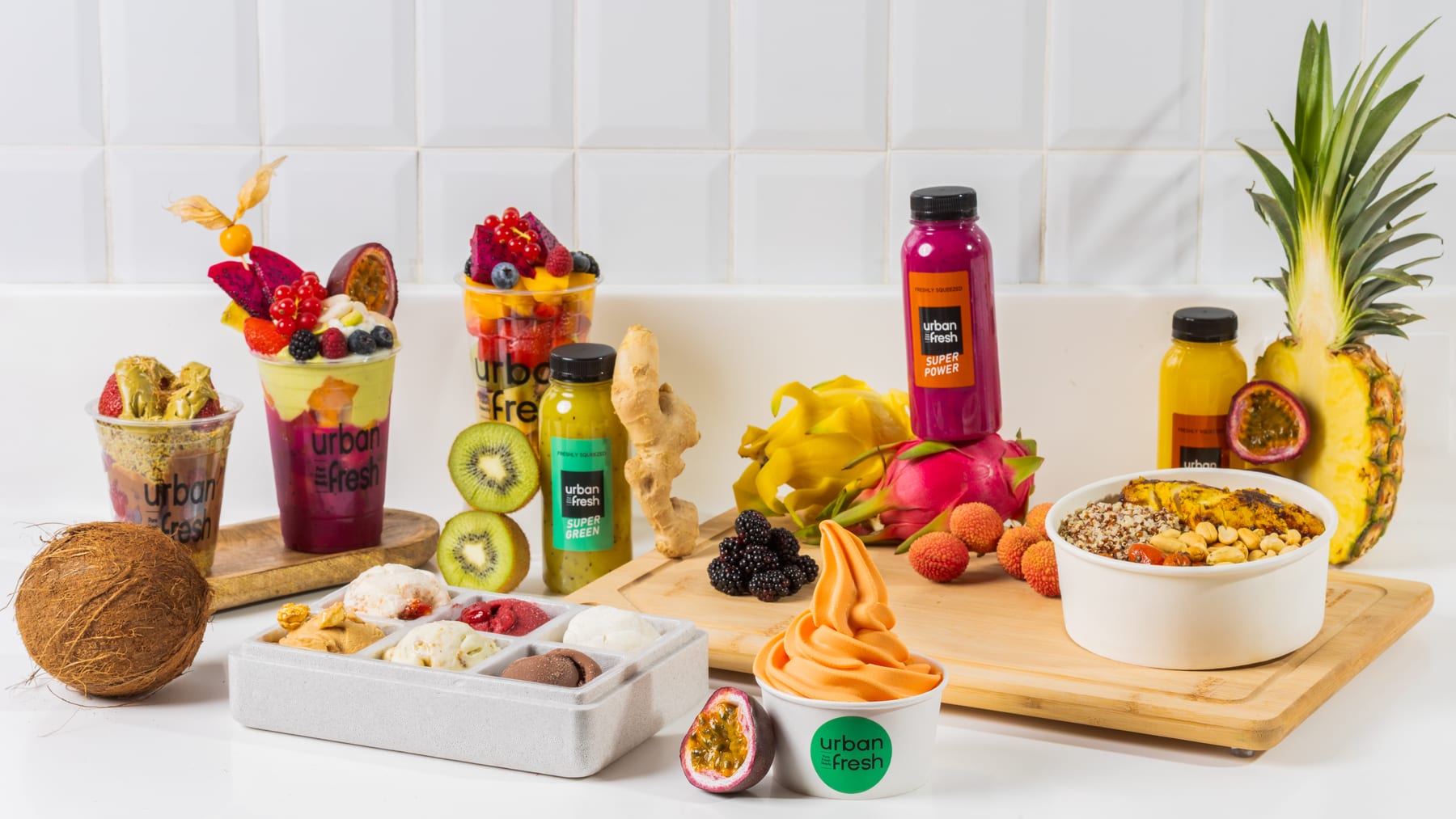 An Urban Fresh spread of smoothie cups, juice bottles, an açai bowl, pineapple, and coconut on white tile